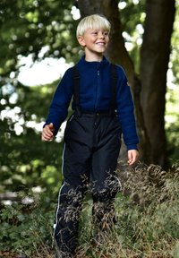 Blaue Fleecejacke mit Reißverschluss, schwarze wasserdichte Latzhose mit verstellbaren Trägern, weißer Seitenbesatz, stehend in einer natürlichen Umgebung zwischen Gras.