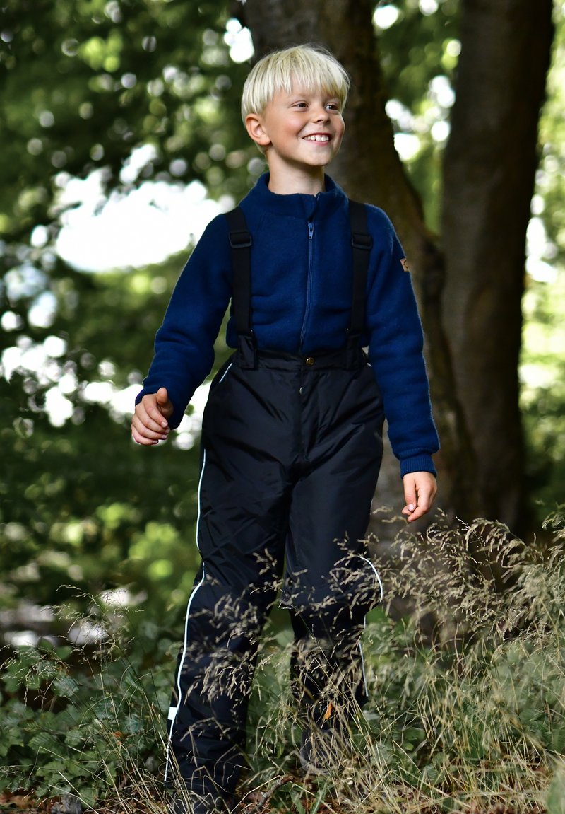 Blaue Fleecejacke mit Reißverschluss, schwarze wasserdichte Latzhose mit verstellbaren Trägern, weißer Seitenbesatz, stehend in einer natürlichen Umgebung zwischen Gras.