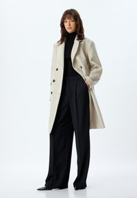 Beige uldfrakke med store mørke knapper, skræddersyet pasform og brede revers, parret med en sort ribbet turtleneck og sorte bukser.