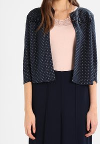 Veste bleu marine à pois avec une texture douce, manches trois-quarts et encolure arrondie, portée par-dessus un haut rose clair.