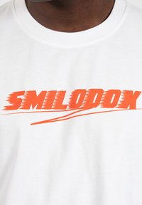 Hvid bomulds t-shirt med en fed orange grafisk tekst "SMILODOX" med en bølget understregning, rund halsudskæring og blød stoftekstur.