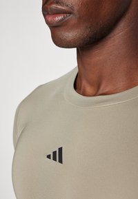 Béžové sportovní tričko s kulatým výstřihem, vyrobené z hladkého materiálu. Na horní levé straně má černé logo. Viditelná texturovaná pleť.