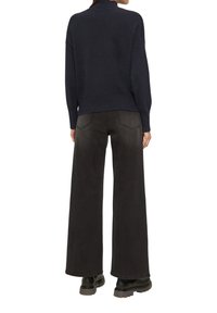 Navy gestrickter Pullover mit hohem Kragen, kombiniert mit weit geschnittenen schwarzen Jeans. Die Jeans haben Gesäßtaschen und eine leichte Textur. Schwarze Stiefel runden das Outfit ab.