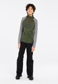 Groene lange mouwen pullover met grijze mouwen, voorzien van een rode rits en zwart logo. Gecombineerd met zwarte broek en stevige zwarte laarzen.