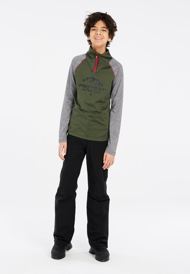 Groene lange mouwen pullover met grijze mouwen, voorzien van een rode rits en zwart logo. Gecombineerd met zwarte broek en stevige zwarte laarzen.