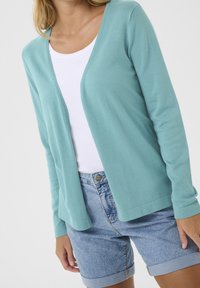 Jasnoniebieski kardigan wykonany z miękkiego materiału, z dekoltem w serek, długimi rękawami i otwartym przodem, zestawiony z szortami jeansowymi.