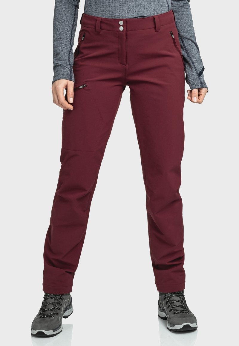 Femme portant un pantalon d'extérieur bordeaux avec poches zippées, des chaussures de randonnée grises et un haut gris à manches longues, debout sur un fond blanc.