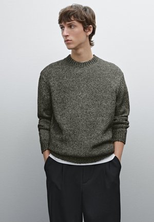 Pull en tricot texturé vert foncé avec un col rond et une coupe légèrement ample, associé à un pantalon sombre, les mains dans les poches.