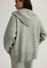 Jasnoszara, oversize, bluza z kapturem z regulowanym sznurkiem, z prążkowanymi mankietami i dołem oraz miękką teksturą materiału. Szwy na plecach.