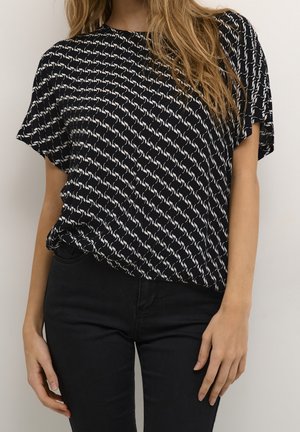 Femme portant un chemisier noir avec un motif abstrait blanc et un pantalon noir, debout devant un fond clair uni.