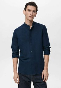 Navy blue button-up shirt made of soft fabric, with a stand collar, long sleeves, and rolled cuffs, paired with dark denim jeans.