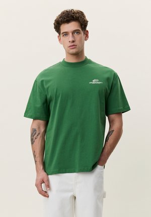 Giovane uomo con capelli ricci che indossa una maglietta verde con piccolo logo bianco e pantaloni bianchi, in piedi con una mano in tasca.