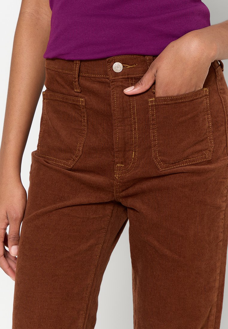 Braune Cordhose mit hoher Taille, zwei vorderen Taschen, gelben Nähten und einem Metallknopfverschluss. Die Textur ist gerippt und weich.