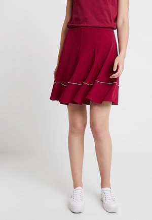 Femme portant une jupe rouge mi-genoux à ourlet froncé et détails de couture blancs, associée à des baskets blanches, debout sur un fond uni.