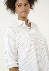 Biała koszula w niebieskie pionowe paski, z guzikiem z przodu, kołnierzykiem i podwiniętymi rękawami. Tkanina wydaje się lekka i gładka. Dwie kieszenie na piersi.