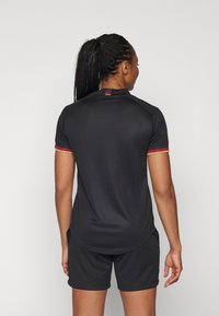 Svart atletisk skjorta med korta ärmar, meshstruktur och röda/guldiga accenter på ärmarna. Matcher med svarta shorts.