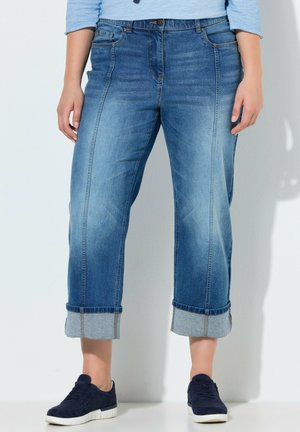 Vrouw met omgeslagen blauwe jeans en marineblauwe sneakers, staand tegen een witte muur.