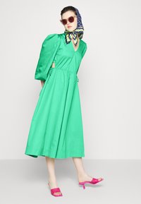 Vestido verde, até aos joelhos, com mangas bufantes e decote em V, apresentando uma cintura ajustável. Combinado com mules de salto rosa e um lenço estampado.