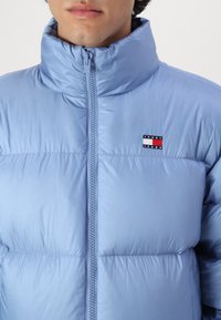 Die hellblaue Pufferjacke besitzt einen hohen Kragen, einen Reißverschluss, eine gesteppten Textur und ein kleines Markenlabel auf der oberen linken Brust.