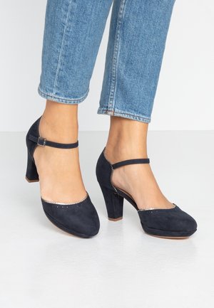 Pieds chaussés de talons Mary Jane en daim bleu avec brides à la cheville, assortis à un jean en denim bleu clair raccourci, sur un sol blanc.