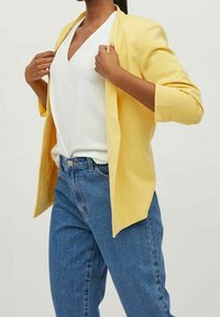 Keltainen bleiseri avonaisella edellä, yhdistetty valkoiseen ribbivartiseen toppiin ja sinisiin korkeavyötäröisiin farkkuihin. Sujuva kangastuntuma, kolmanneksen pituiset hihat.