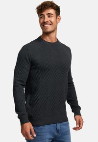 Mørkegrå strikket sweater med et forhøjet fiskebensmønster, rund halsudskæring og ribbede manchetter og nederkant. Båret med blå jeans.