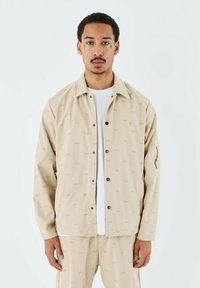 Chaqueta beige de botón con un tejido estampado que presenta las letras "PXP". Incluye un bolsillo en el pecho y está hecha de un material ligero.