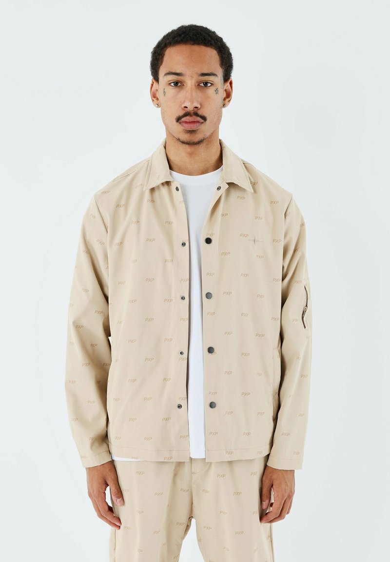 Chaqueta beige de botón con un tejido estampado que presenta las letras "PXP". Incluye un bolsillo en el pecho y está hecha de un material ligero.