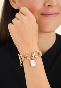 Gouden horloge met een rechthoekige witte wijzerplaat, geometrische schakels en een toggle sluiting, passend bij een gladde huidtextuur.