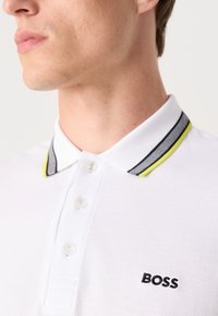 Hvit polo-skjorte med en svart og gulstripet krage, med tre knapper og et brodert "BOSS"-merke på brystet.