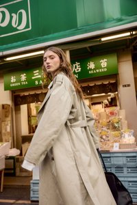 Gai krēmkrāsas tranče palto ar pārspīlētām piedurknēm un jostu viduklim, saskaņots ar melnu somu. Rāda gludu tekstūru tirgus fona priekšā.