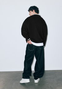 Zwarte oversized trui met witte accenten, gecombineerd met losse donkere denim jeans en witte sneakers, tegen een effen witte achtergrond.