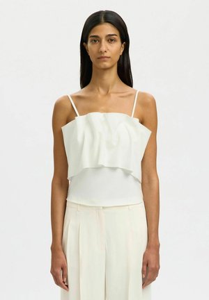 Kobieta o długich ciemnych włosach, ubrana w białą falbaniaste bezrękawnik i kremowe plisowane spodnie, stojąca na jednolitym tle.