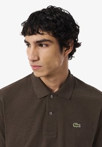 Giovane uomo con capelli neri e ricci che indossa una polo marrone con un logo di coccodrillo verde, guarda a sinistra su uno sfondo bianco.