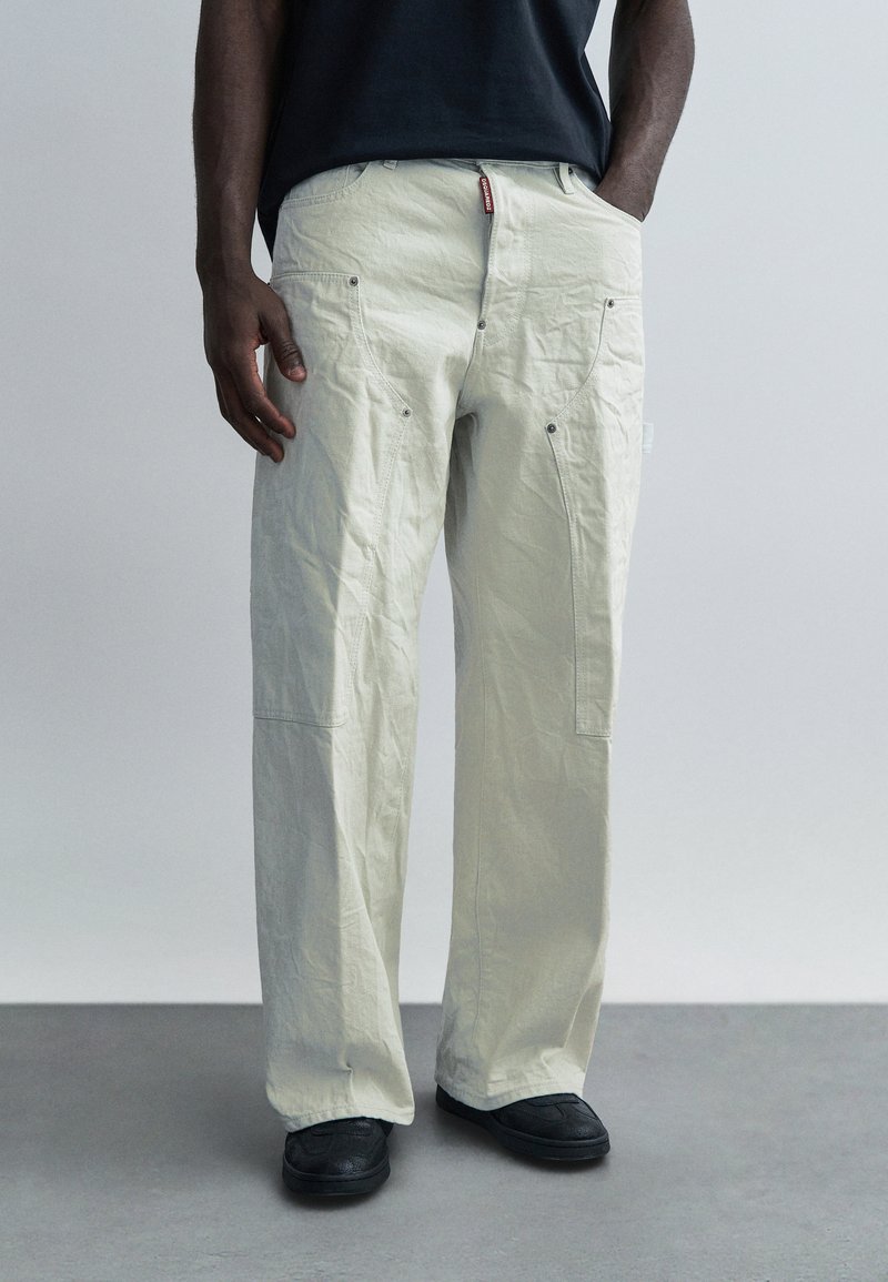 Pantalones beige claro de pierna ancha con textura arrugada, que cuentan con bolsillos laterales, remaches de metal y una etiqueta en la cintura.