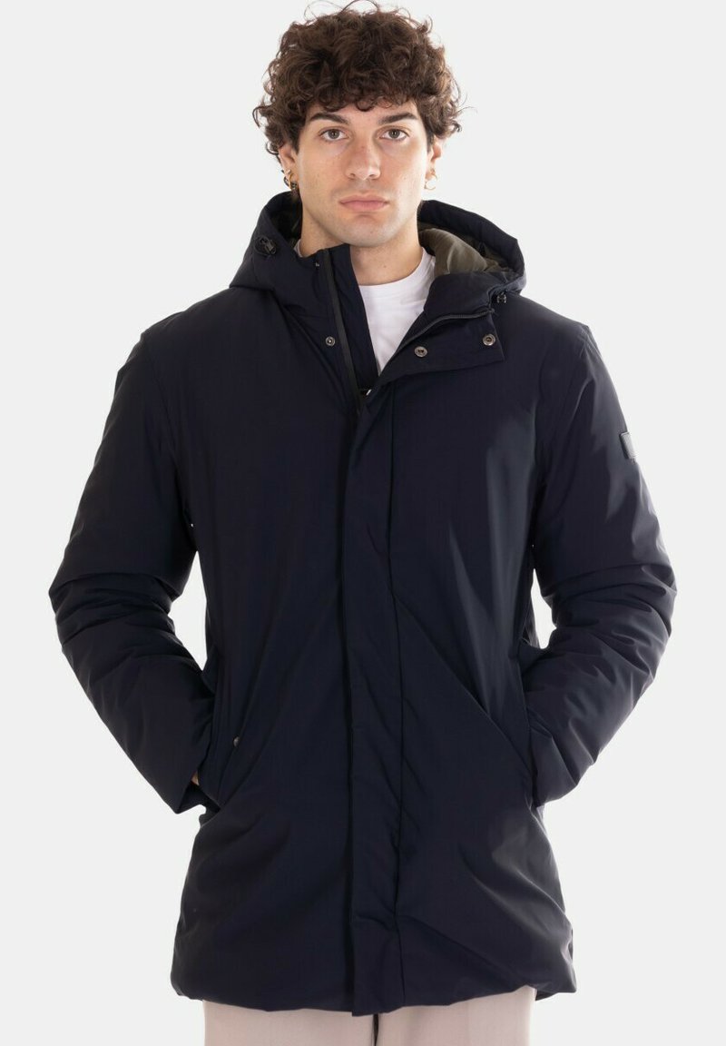 Giovane uomo che indossa una giacca invernale nera con cappuccio, con le mani in tasca, in piedi davanti a uno sfondo chiaro e uniforme.