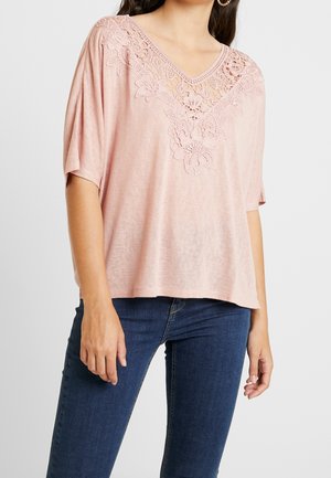 Jasnoróżowy top z krótkim rękawem, z dekoltem w serek i misternymi koronkowymi kwiatowymi zdobieniami wzdłuż linii szyi. Miękki materiał o luźnym kroju.