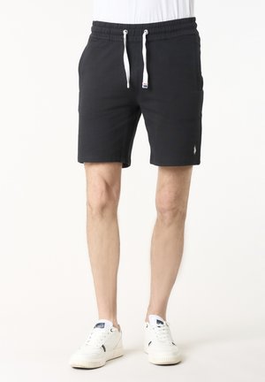 Uomo che indossa pantaloncini neri con coulisse e sneakers bianche, in piedi davanti a uno sfondo chiaro e semplice.