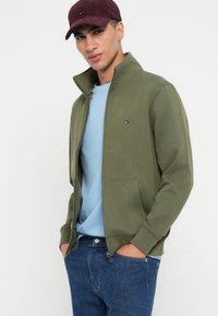 Ελαιόχρωμο ζακέτα με φερμουάρ και ψηλό γιακά, δύο μπροστινές τσέπες και ένα μικρό λογότυπο. Φορεμένο πάνω από ένα ανοιχτό μπλε μπλουζάκι και συνδυασμένο με μπλε τζιν.