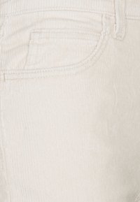 Tissu en velours côtelé blanc avec des côtes verticales, comportant une poche avant avec des accents de couture et une fermeture à glissière.