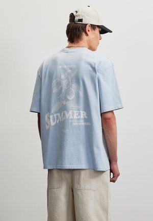 Hombre con camiseta de gran tamaño azul claro con gráfico botánico y texto "SUMMER", pantalones cortos beige y gorra de béisbol blanca visto de lado.