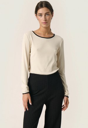 Haut crème à manches longues avec un col noir, associé à un pantalon noir à côtes. Le tissu semble lisse, avec une silhouette ajustée.