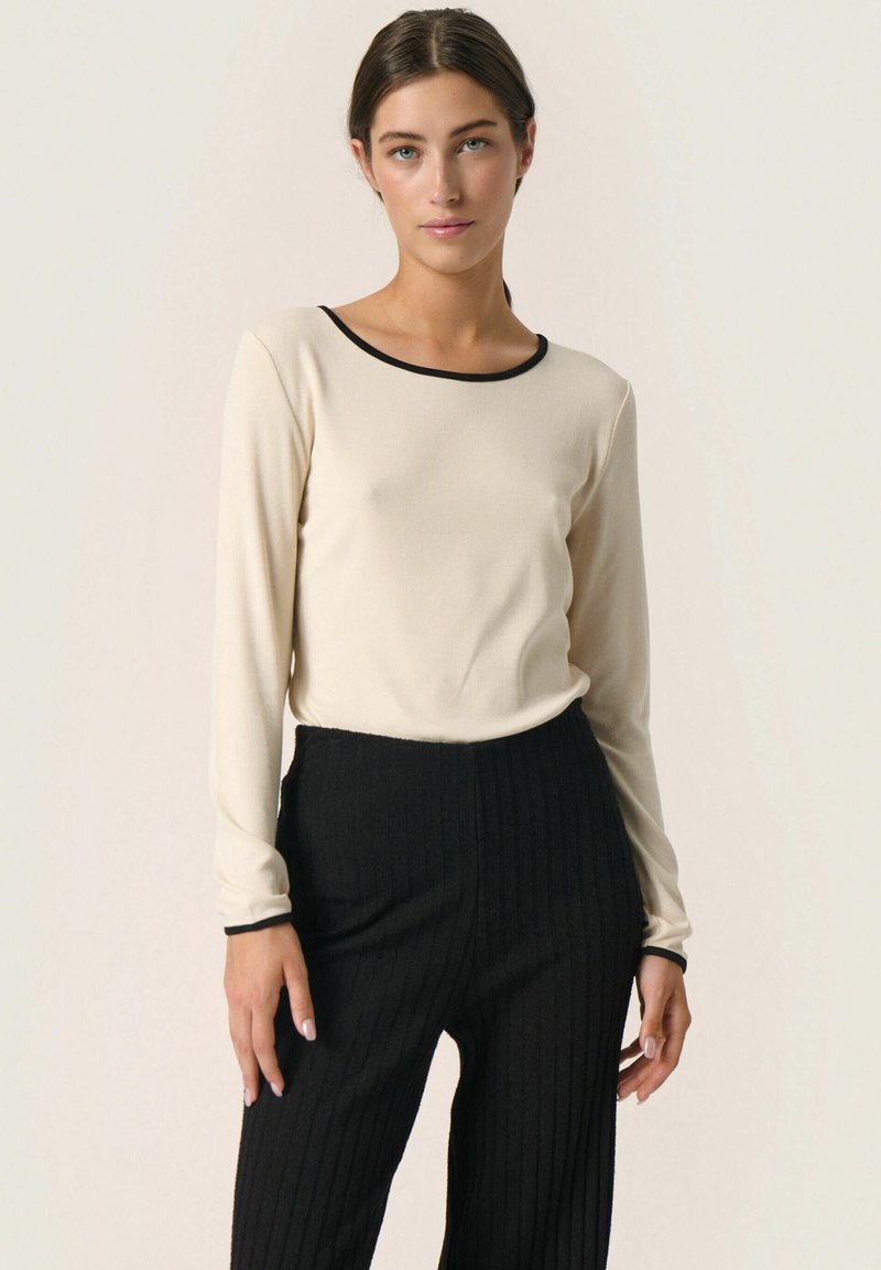 Top a maniche lunghe crema con collo nero, abbinato a pantaloni neri a coste. Il tessuto appare liscio, con una silhouette aderente.