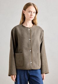 Brun houndstoothjakke med rund hals, front snaplukninger, to store lommer og brede ærmer. Tekstureret stof med kontrasterende kant.