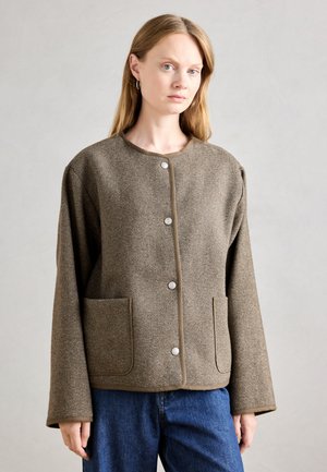 Brun houndstoothjakke med rund hals, front snaplukninger, to store lommer og brede ærmer. Tekstureret stof med kontrasterende kant.