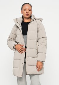 Manteau matelassé beige clair avec capuche, présentant une texture matelassée, une fermeture éclair et des poches latérales. Accents en matériel doré.
