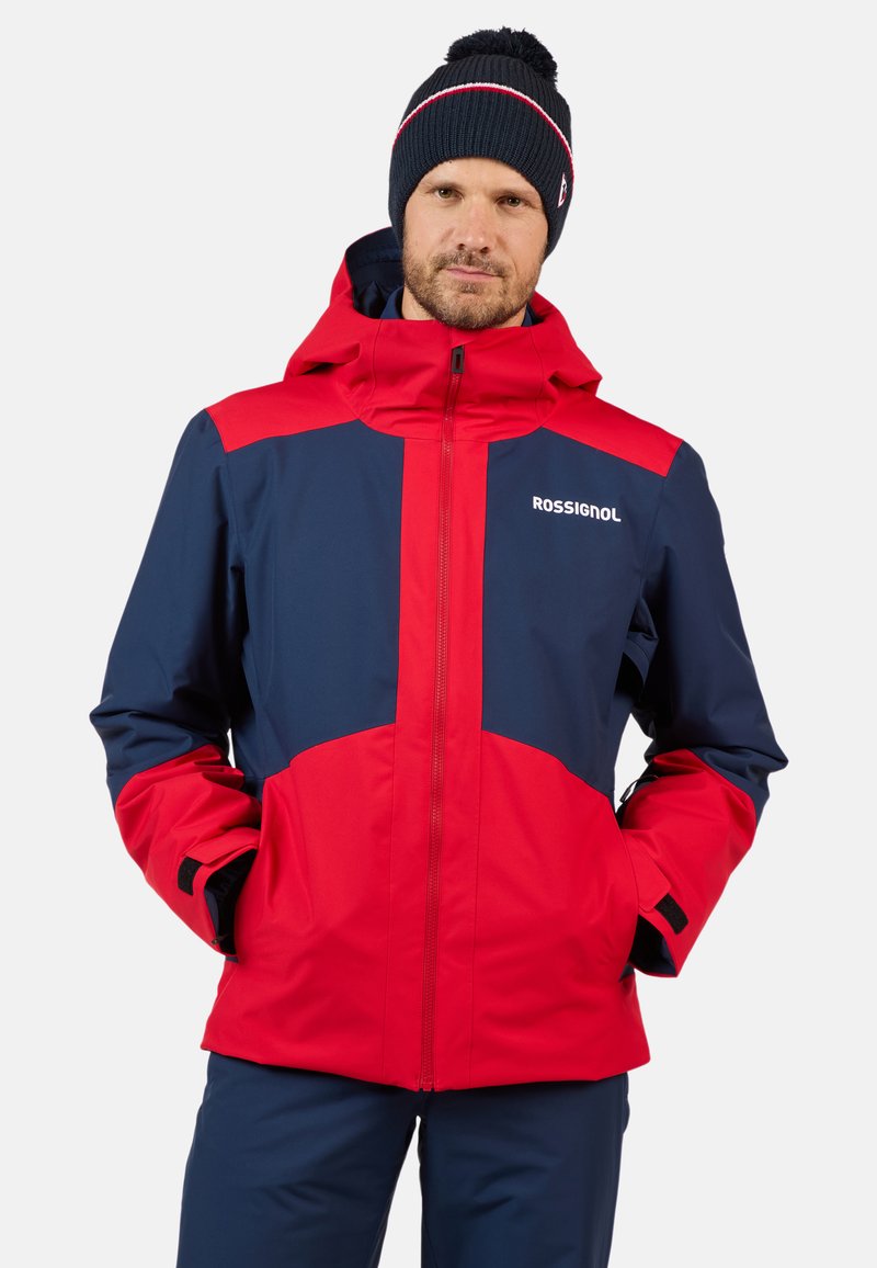 Giacca impermeabile rossa e blu con chiusura a zip, cappuccio regolabile e polsini in Velcro. Caratterizzata da colori a contrasto e un logo sul petto.