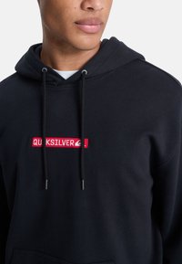 Sudadera de algodón negra con un parche de logo bordado en rojo en el pecho, equipada con capucha ajustable y un bolsillo canguro.