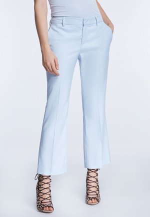 Femme portant un pantalon droit bleu clair avec les mains dans les poches et des sandales à talons hauts à lacets imprimé léopard sur un fond uni.