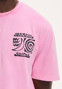 Šviesiai rožinė medvilninė marškinėliai su juodu grafiniu dizainu, kuriame yra abstrakčių formų ir tekstas "Mezcal Paloma" kairėje krūtinės pusėje.